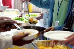 Large team of volunteers serve healthy vegetable soup and salad, potato chips and other food items to homeless people. View is of hands serving others.